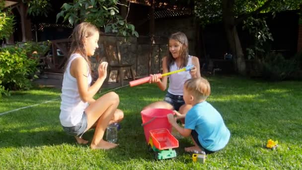 Slow motion footage of little toddler boy filling water gun from the bucket and shooting in two teenage girls at house backyard — Stock Video