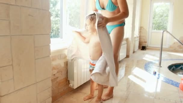 4k felvételeket mosolygó fiatal anya szárítás és felmelegedés neki kisgyermek fia nagy törülközővel osztály után a medencében — Stock videók