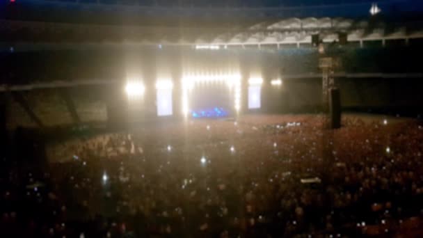 Fora de foco vídeo de cena brilhantemente iluminada em concerto no grande estádio. Multidão está dançando e acenando com telefones celulares — Vídeo de Stock