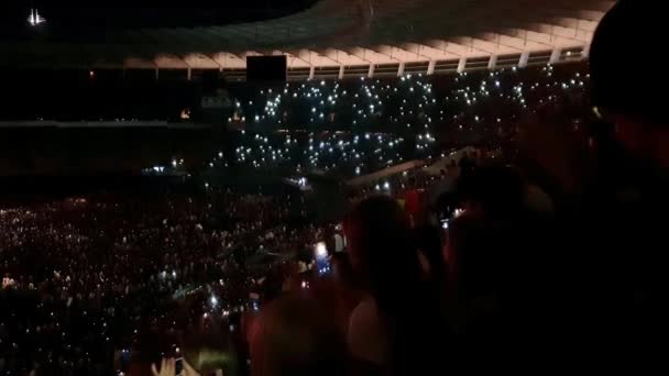 4k fuera de foco de vídeo de personas en tribunas de estadio saludando con teléfonos inteligentes y antorchas de luz mientras escucha música en concierto de rock por la noche. Multitud de fanáticos del grupo de rock en la arena deportiva por la noche — Vídeos de Stock