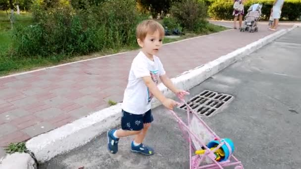 Imágenes de 4k del adorable niño pequeño caminando por la calle con un cochecito de juguete para muñeca. Chico jugando con juguetes para chicas. Bebé activo — Vídeo de stock