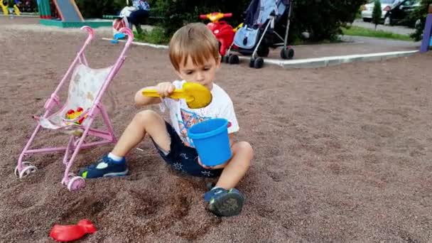 4k镜头的幼儿男孩挖沙与勺子，并倒入塑料玩具桶 — 图库视频影像