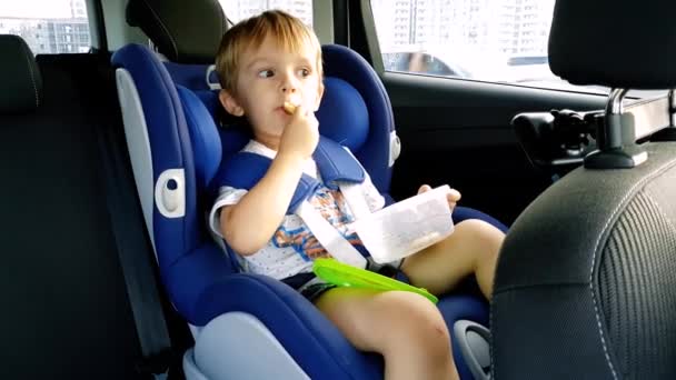 4K πλάνα από 3 χρόνια νήπιο παιδί κάθονται στο παιδικό κάθισμα ασφαλείας του αυτοκινήτου και τρώγοντας — Αρχείο Βίντεο