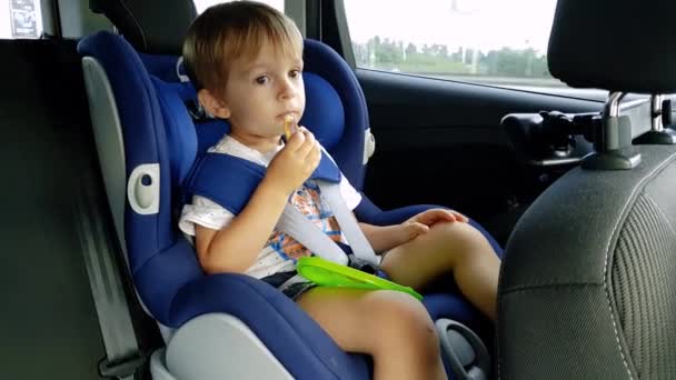 4k видео маленького мальчика ест во время езды в машине. Ребенок сидит на автокресле и ест печенье — стоковое видео