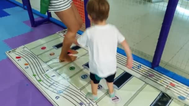 4K bilder av glada glada småbarn pojke hoppar och leker med Big piano på golvet i nöjesparken. Stepping på stora tangentbord av interaktiva elektroniska piano. — Stockvideo