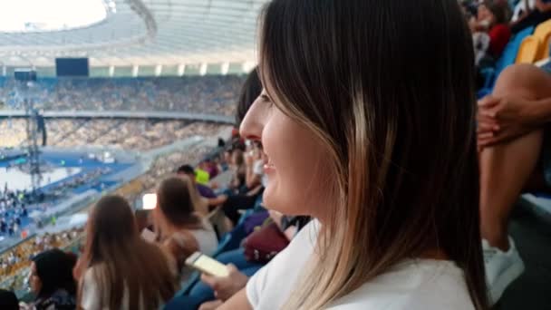 4k крупным планом видео красивой улыбающейся молодой женщины с длинными волосами, сидящей на трибунах большого стадиона и смотрящей футбольный матч — стоковое видео