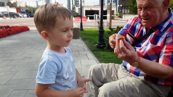 4k video of senior grandfather playing with his toddler grandson with toy on bench at park — Stock Video