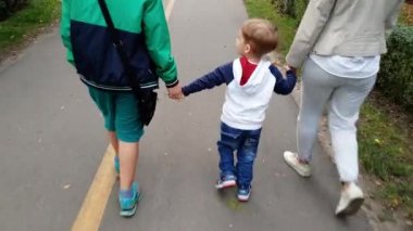 Parkta yürüyen annesi ve ağabeyini elle tutan sevimli gülümseyen küçük çocuğun 4k videosu