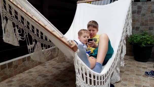 4k-beelden van ouderling Boy met toddeler swingen in hangmat en met behulp van slimme telefoon — Stockvideo