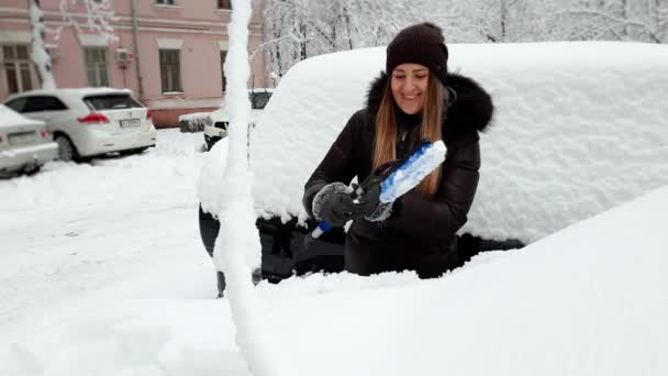 4k видео улыбающейся женщины-водителя, убирающей снег из своей машины утром после снежной бури — стоковое видео