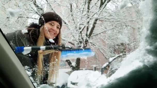 ब्रश के साथ बर्फ से कार विंडस्क्रीन सफाई करने वाली सुंदर मुस्कुराते हुए युवा महिला का 4k दृश्य वीडियो — स्टॉक वीडियो