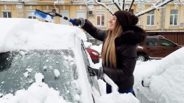 Wideo 4K uśmiechniętej kobiecej kierowcy usuwający śnieg z samochodu o poranku po burzy śnieżnej — Wideo stockowe