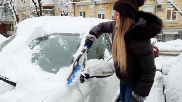 4k відео красивої усміхненої молодої жінки, що прибирає сніг з її машини з пензлем на парковці — стокове відео