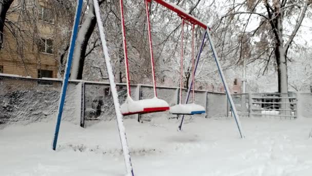 Imágenes de 4k de columpios vacíos en el patio cubierto de nieve balanceándose por el viento — Vídeo de stock