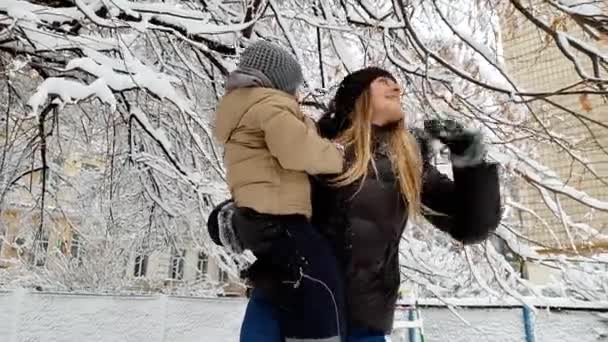 4k视频的年轻母亲抱着她蹒跚学步的儿子和摇动的树枝覆盖着雪。家庭在冬季公园玩得开心 — 图库视频影像
