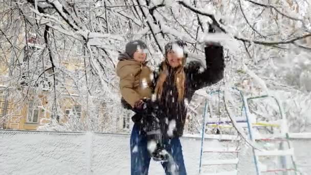 4k видео счастливой улыбающейся молодой женщины, которая держит своего маленького мальчика и играет со снегом в зимнем парке — стоковое видео