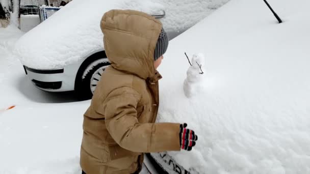 Imágenes de 4k del niño mirando al muñeco de nieve pequeño en el capó del coche — Vídeos de Stock