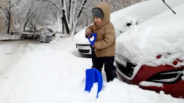 4k видео счастливого улыбающегося мальчика, копающего снежные заносы на автостоянке с большой снежной лопатой — стоковое видео