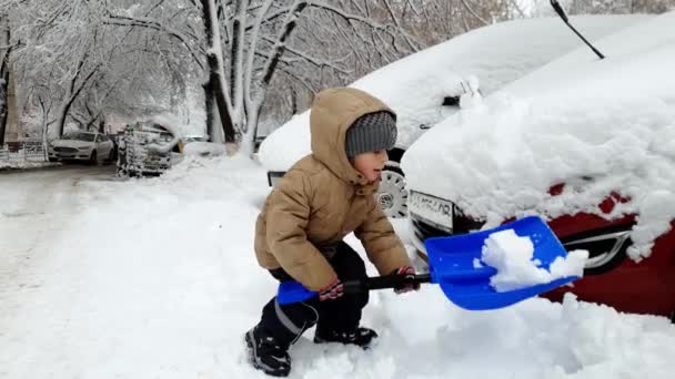 Imágenes de 4k de un niño pequeño aserrando un auto atrapado en la nieve después de la tormenta de nieve. Niño cavando nieve con pala — Vídeo de stock