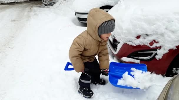4k видео маленького 3-летнего мальчика, копающего снег на автостоянке лопатой — стоковое видео