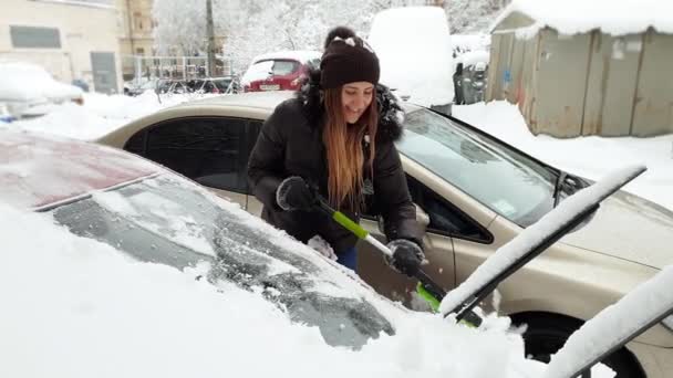 4k video van mooie glimlachende jonge vrouw het schoonmaken van haar auto uit sneeuw met telescopische borstel — Stockvideo