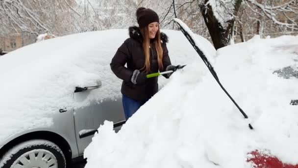 4k video de hermosa mujer joven sonriente quitando la nieve de su coche con brish en el estacionamiento — Vídeos de Stock