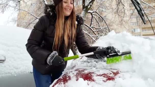 4K video av vackra leende ung kvinna rengöring bil vindruta från snö med borste — Stockvideo