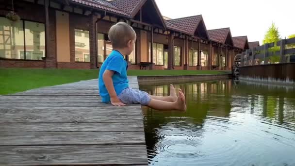 4k vídeo de chererful sorrindo criança menino sentado na margem do rio e segurando os pés na água de pequeno canal na cidade europeia — Vídeo de Stock