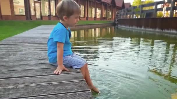 Filmagem 4K de um adorável menino criança sentado na margem do rio e mergulhando os pés na água. Crianças movendo as pernas e salpicando água — Vídeo de Stock