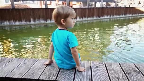 4k video of chererful smiling toddler boy sitting on the riverbank and holding feet in water of small canal at european town — Stock Video