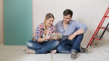 Yeni evlerinin zemininde tadilatta oturan ve tablet bilgisayar kullanarak internetten mobilya seçen genç çiftin 4k görüntüleri