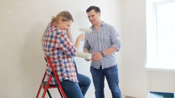 Glückliches junges Paar, das Spaß hat und sich bei Renovierungsarbeiten in ihrem neuen Haus mit Farbwalzen schlägt — Stockvideo