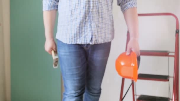 Primer plano video en cámara lenta de un joven trabajador masculino sosteniendo un sombrero rojo y un martillo en la mano caminando en casa en construcción — Vídeos de Stock