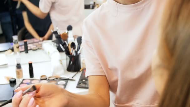 Slow motion video of young beautiful fashion model sitting next to big mirror with bulbs while professional makeup artist is applying makeup on her face — Stock Video