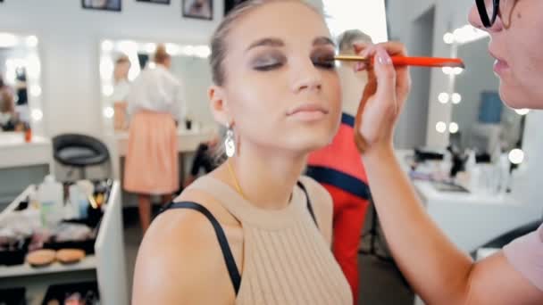 Vidéo au ralenti de la jeune belle mannequin assise à côté d'un grand miroir avec des ampoules tandis que le maquilleur professionnel applique du maquillage sur son visage — Video