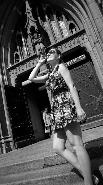 Black and white portrait of beautiful smiling young woman in short dress and sunglasses posing against old catholic church — Stock Photo, Image