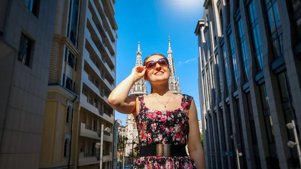 Retrato de hermosa joven sonriente en gafas de sol posando contra edificio moderno y antigua catedral católica en la ciudad europea — Foto de Stock