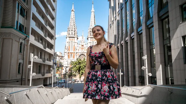 Portret van mooie glimlachende jonge vrouw in zonnebril poseren tegen modern gebouw en oude katholieke kathedraal in de Europese stad — Stockfoto