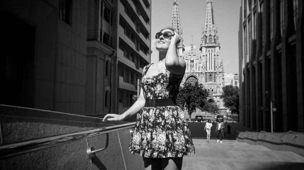 Black and white portrait of beautiful smiling young woman in short dress walking on streets old old european town with modern office building and ancient churches — Stock Photo, Image