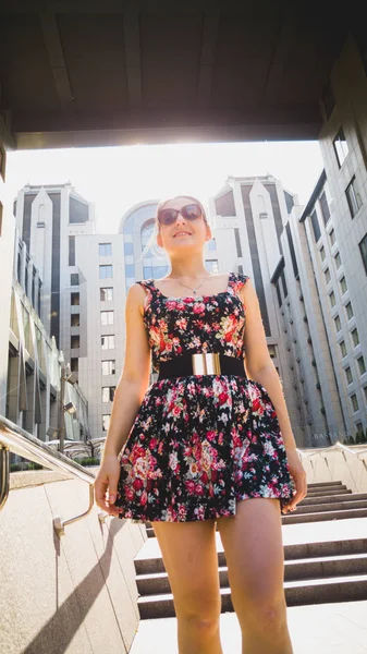 Portrait of sexy young woman in short dress posing against modern building made of concrete and glass in sun light rays — Stock Photo, Image