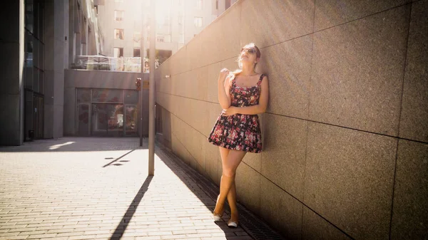 Şehir caddesindeki taş duvara yaslanmış ve kameraya bakan kısa elbiseli güzel genç kadının portresi — Stok fotoğraf