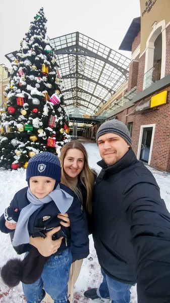 Selfie-Porträt einer glücklichen jungen Familie mit kleinem Jungen, der im Winter auf der Straße der Altstadt vor einem großen geschmückten Weihnachtsbaum posiert — Stockfoto