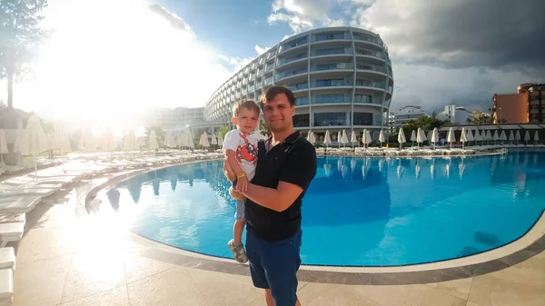 Portrait of happy young father hugging his toddler son against big swimming pool at hotel resort. Family relaxing on summer beach holiday vacation — Stock Photo, Image