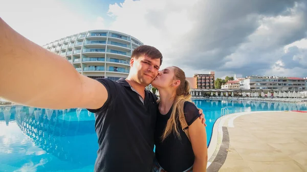 Beautiful young woman kissing her husband while he is making selfie on smartphone against big outdoor swimming pool at hotel resort. Family relaxing on summer beach holiday vacation — Stock Photo, Image