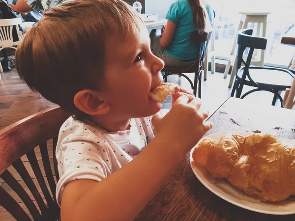 Toned closeup portrait of adorable toddler boy eating freshly baked croissant in the bakery — Stock Photo, Image