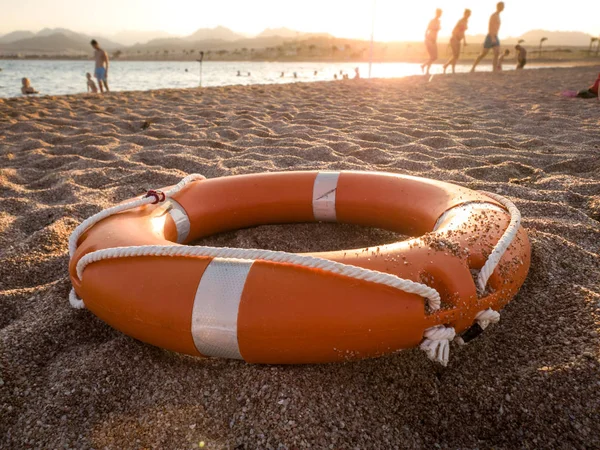 Foto de primer plano del anillo de plástico rojo para salvar la vida en la arena de la playa del mar contra la hermosa puesta de sol sobre el océano —  Fotos de Stock