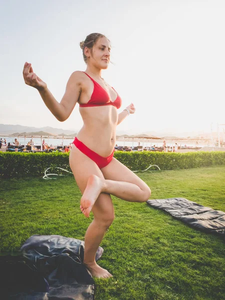 Toned image of young slim woman wearing red bikini doing yoga exercises on the grass next to the sea beach — Stock Photo, Image