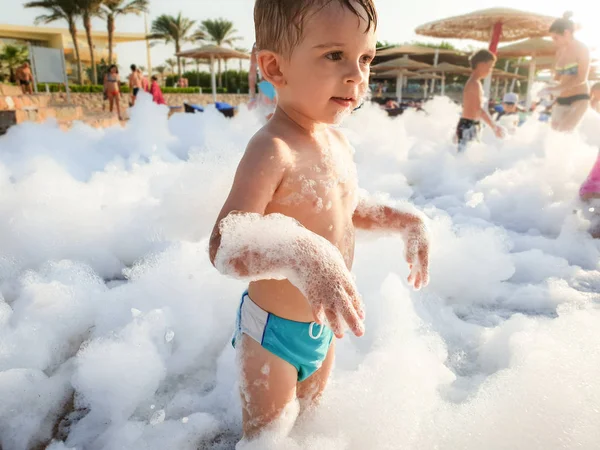 Забавный портрет маленького мальчика, покрытого мыльной пеной, играющего на пляже во время дискотеки — стоковое фото