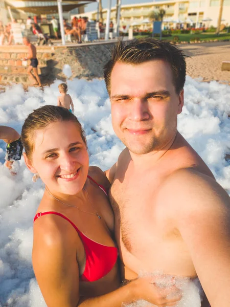 Toned portrait of happy young couple making selfie portrait of the soap foam disco party at sea beach — Stock Photo, Image