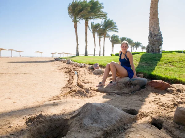 Beautiful young woman with long hair sitting under big palm tree at sea beach — Stock Photo, Image